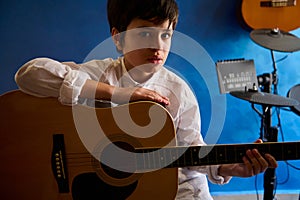 Handsome teenage boy playing guitar in the modern music studio, looking confidently at camera, sitting against a blue