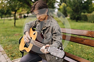 Handsome teenage playing acoustic guitar outdoor in Autumn time. Boy sitting on bench and playing music