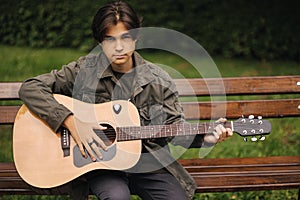 Handsome teenage playing acoustic guitar outdoor in Autumn time. Boy sitting on bench and playing music