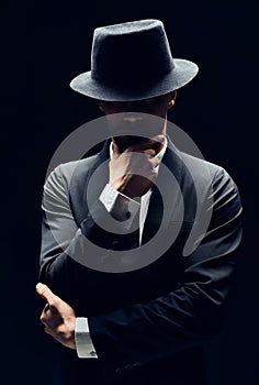 Handsome thoughtful man in black suit and hat touching chin on dark background