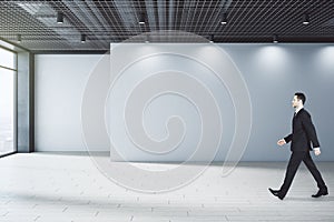 Handsome thoughtful young european businessman walking in concrete interior with empty mock up place on wall, city view and