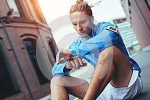 Handsome tired athlete in blue sitting on the street and checking running results, resting after jogging