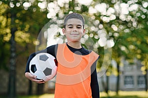 Handsome tired boy in football uniform with ball after training on the stadium in the urban green park on summer day.