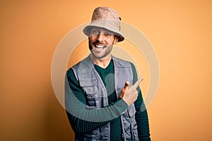 Handsome tourist man with beard on vacation wearing explorer hat over yellow background cheerful with a smile on face pointing