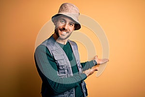 Handsome tourist man with beard on vacation wearing explorer hat over yellow background Inviting to enter smiling natural with