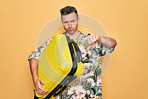 Handsome tourist man with blue eyes on vacation holding suitcase over yellow background with angry face, negative sign showing