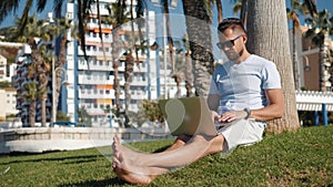 Handsome tourist man student in glasses hipster using laptop in park on a summers day sitting on grass city park street