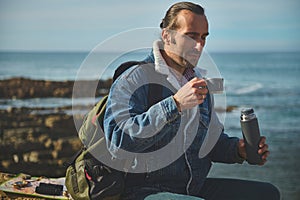 Handsome traveler man holds a thermos flask and drinks tea or coffee from stainless steel mug, sitting on a rock by sea