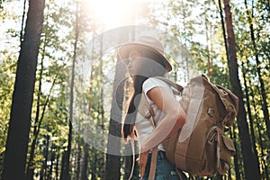 Handsome traveler woman with backpack and hat standing in forest. Young hipster girl walking among trees on sunset