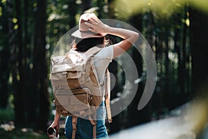 Handsome traveler woman with backpack and hat standing in forest. Young hipster girl walking among trees on sunset photo