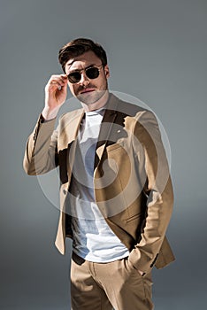 Handsome trendy man in beige suite and sunglasses looking at camera on grey.