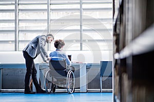 Handsome university professor helping student in wheelchair, showing him book for his thesis in library.