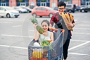 Handsome vietnamese man holds paper bags with food pushing in front of him shopping trolley with his happy beautiful