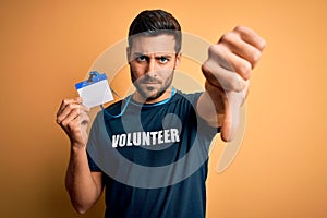 Handsome volunteer man with beard holding id card identification over yellow background with angry face, negative sign showing