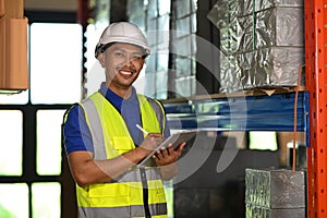 Handsome warehouse worker wearing security vest using digital tablet while standing near the stack of cardboard boxes