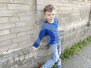 A handsome white boy in a blue sweater or longsleeve is standing against the wall and smiling. Child on a brick wall background.