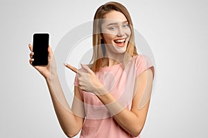 Handsome woman with index finger shows blank phone screen where you can place your ad. Positive young girl smiles snow-white smile