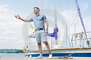 Handsome yachtsman walking at river pier and welcomes visitors. Young businessman invites to his boat for yacht adventure.