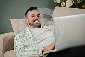 Handsome Young Adult Man Using Laptop While Sitting On Couch At Home. Smiley Millennial Guy Working Remotely On Computer