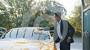 Handsome young African American businessman cleaning his car windscreen with yellow sponge and soap foam outdoors at car