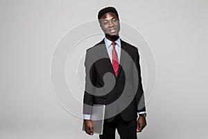 Handsome young African man carrying laptop and looking at camera while standing against grey background