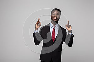 Handsome young African man in suit pointing up and smiling while standing against grey background