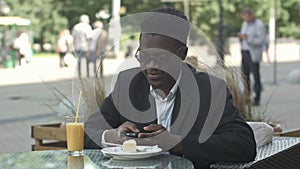 Handsome young afro american businessman using smart phone, messaging his girlfriend, eating at cafe