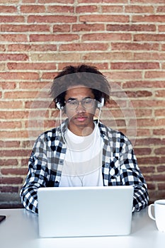 Handsome young afro-american man with eyeglasses listening to music with headphones while using his laptop at home