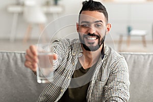 Handsome young Arab guy holding glass of fresh clear water and smiling at camera at home