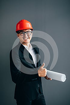 Handsome young architect in suit and protective helmet is showing with blueprint Ok sign, looking at camera and smiling, on gray b