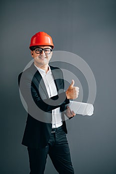 Handsome young architect in suit and protective helmet is showing with blueprint Ok sign, looking at camera and smiling, on gray b