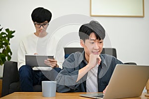 A handsome young Asian male college student using a laptop in the living room with his friend