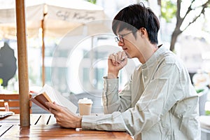 A handsome young Asian man focusing on reading a book while relaxing at a cafe