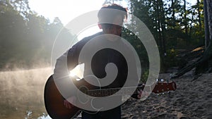 Handsome young asian man playing guitar at coast of forest river at sunrise. Dawn misty fog at background