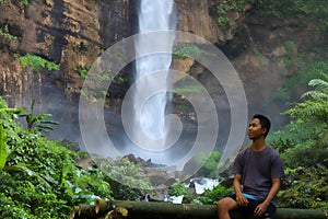 Handsome young asian man sitting on the bamboo bench looking at water in waterfall in sunny day. space for text