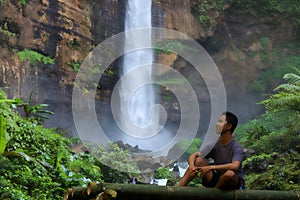Handsome young asian man sitting on the bamboo bench looking at water in waterfall in sunny day. space for text