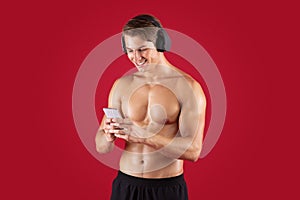 Handsome young athlete with bare torso listening to music in headphones, choosing song on cellphone over red background
