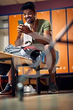 Handsome young athletic man looking joyful looking at smartphone typing a message, taking a break during workout in locker room