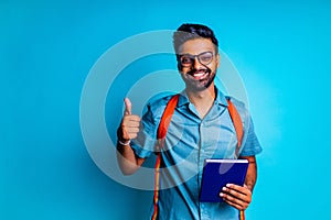 handsome young bearbed indian man with eye glasses in blue cotton t-shirt with orange rainbow backpack i thumbs up n