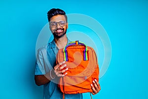 handsome young bearbed indian man with eye glasses in blue cotton t-shirt with orange rainbow backpack in studio