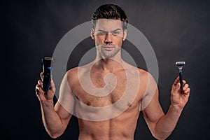Handsome young bearded man isolated. Portrait of topless muscular man is standing on gray background with trimmer in one hand and