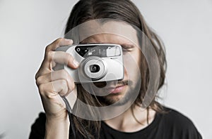 Handsome young bearded man with a long hair and in a black shirt holding vintage old-fashioned film camera on a white