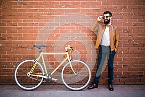 Handsome young bearded man while standing near his bicycle against the brick wall