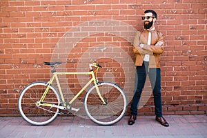 Handsome young bearded man while standing near his bicycle against the brick wall