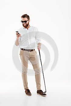 Handsome young bearded man standing over white wall with golfstick