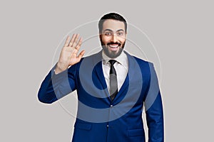 Handsome young bearded man standing waving hand, looking at camera with engaging toothy smile.