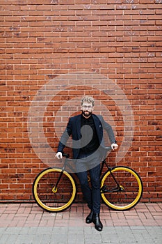 Handsome young bearded man in suit and glasses standing near his bicycle against the brick wall outdoors