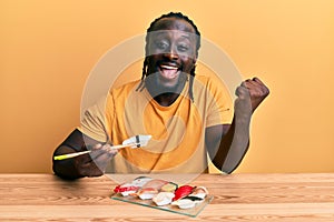 Handsome young black man eating sushi sitting on the table screaming proud, celebrating victory and success very excited with