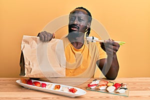 Handsome young black man eating take away sushi sitting on the table clueless and confused expression