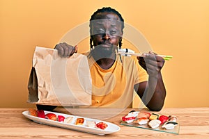 Handsome young black man eating take away sushi sitting on the table puffing cheeks with funny face